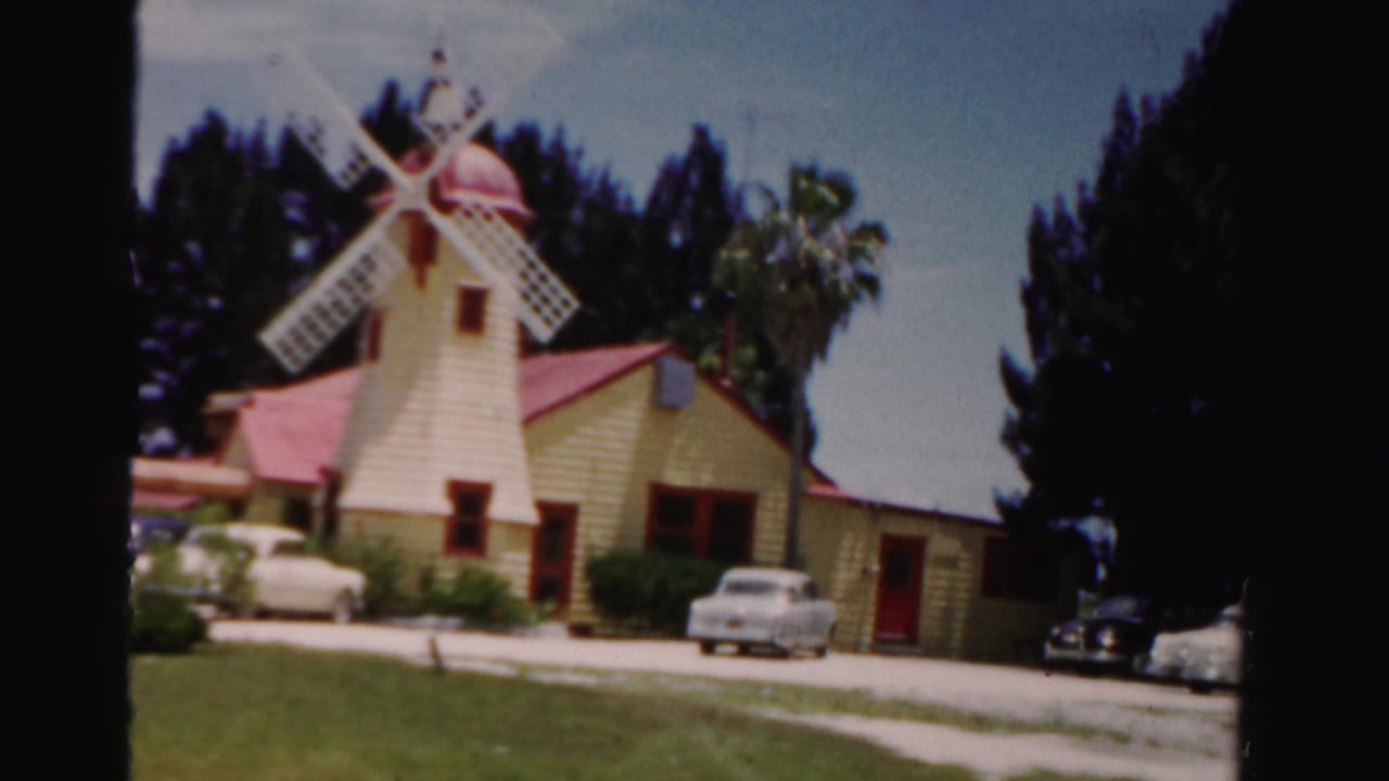 1955 Dade County Florida home-movie frame showing a yellow tourist-attraction building with a windmill, lawn, parked cars, and palms.