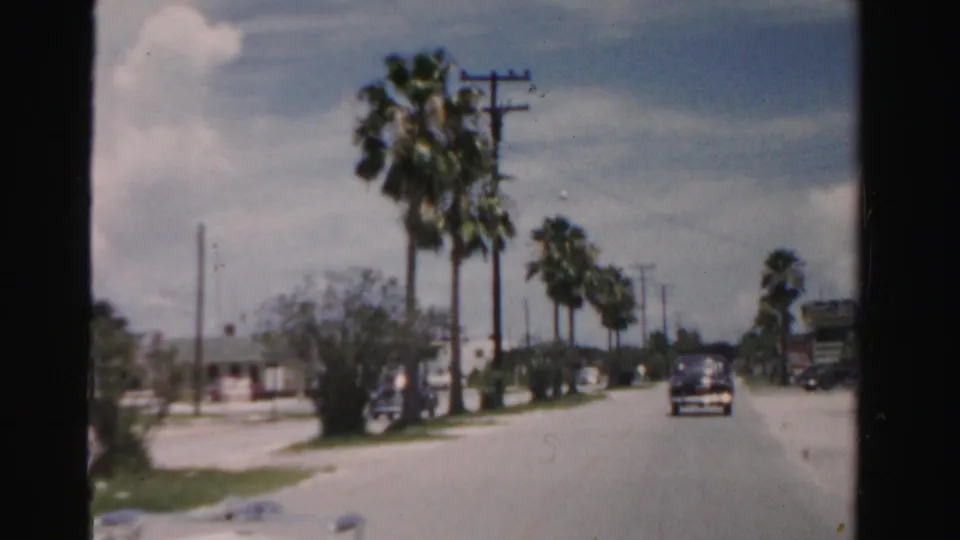 1955 Dade County Florida moving-vehicle view of a quiet road with palms, utility poles, houses, a dark car, and a larger vehicle.