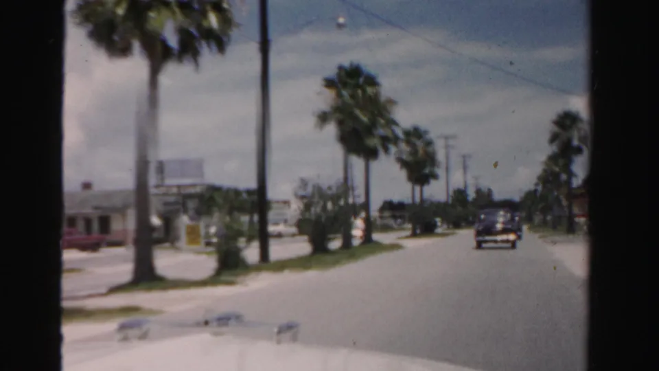 1955 Dade County home-movie view from a moving car on a palm-lined road with a dark automobile ahead and low roadside buildings nearby.