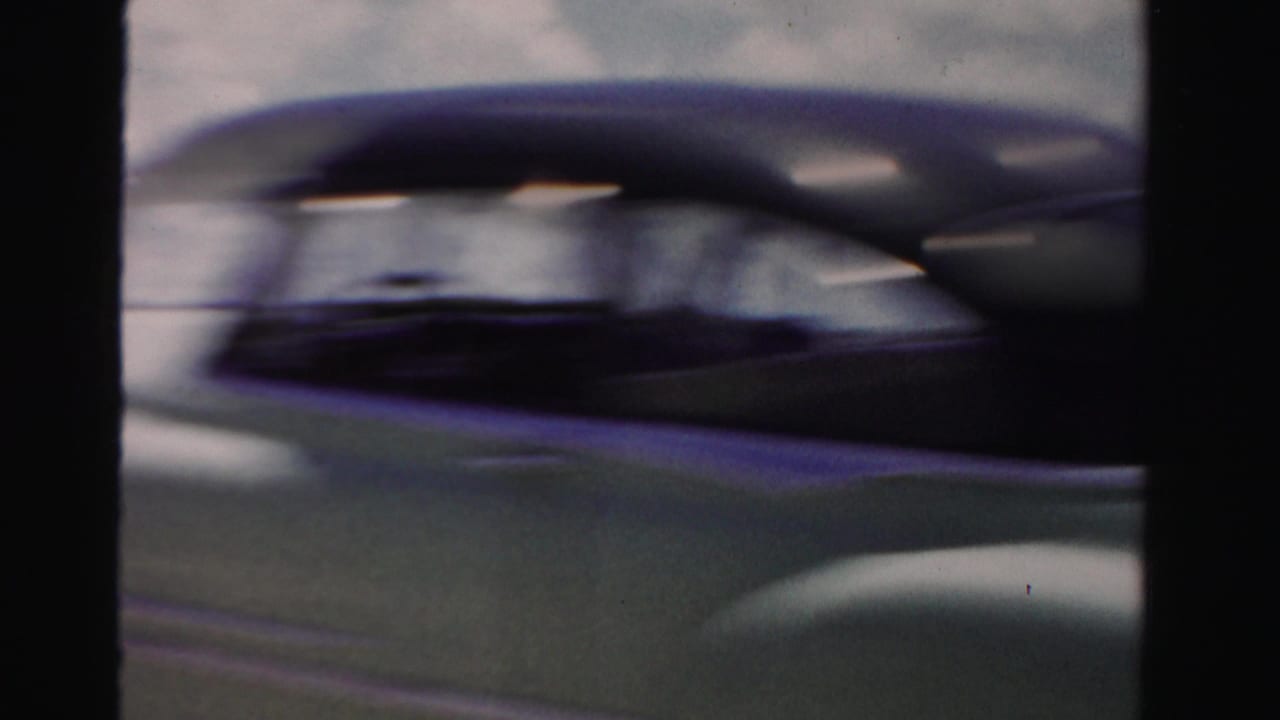A motion-blurred mid-century car passes close beside the filming vehicle, emphasizing the moving home-movie perspective on a Florida road.