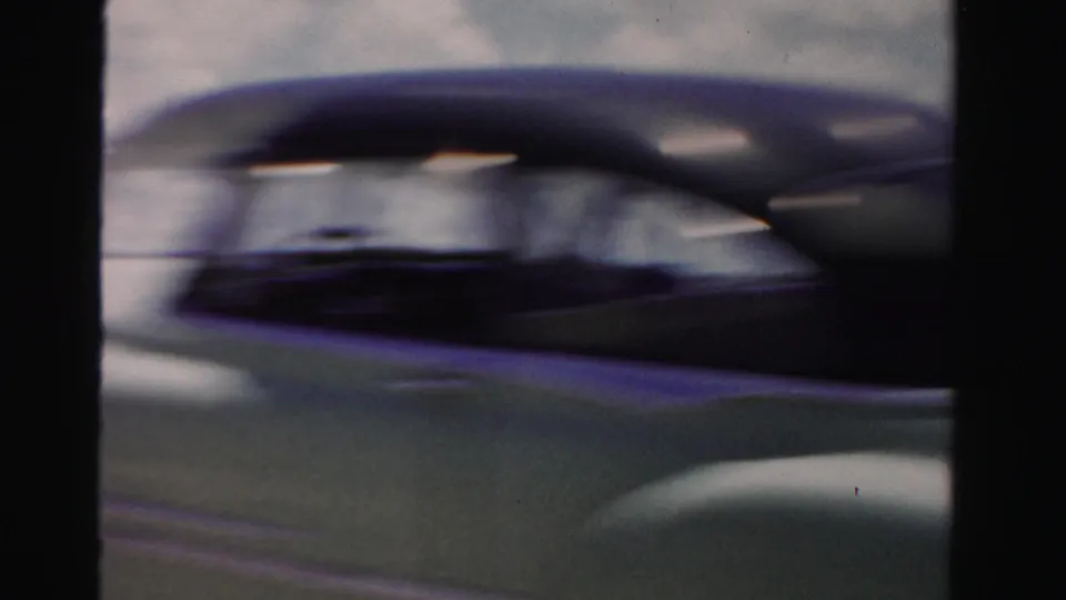 A motion-blurred mid-century car passes close beside the filming vehicle, emphasizing the moving home-movie perspective on a Florida road.