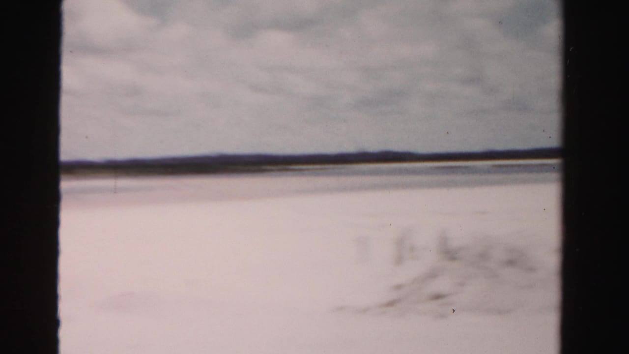 A brief Dade County coastal view from the moving vehicle shows pale beach or waterfront ground, cloudy sky, and a low distant horizon.