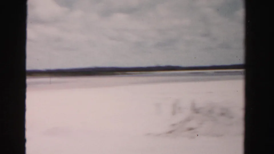 A brief Dade County coastal view from the moving vehicle shows pale beach or waterfront ground, cloudy sky, and a low distant horizon.