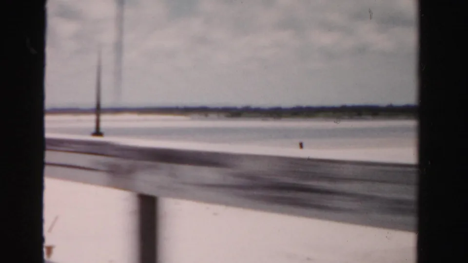 1955 Dade County, Florida travel frame with a lamppost, bridge barrier, open water, and far shoreline.