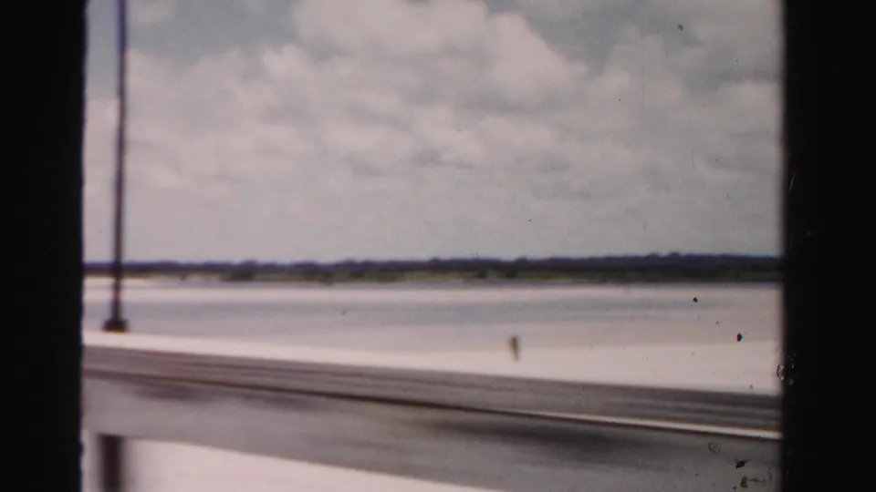 1955 Dade County, Florida home-movie frame with lampposts, bridge rail, water, and distant shoreline.