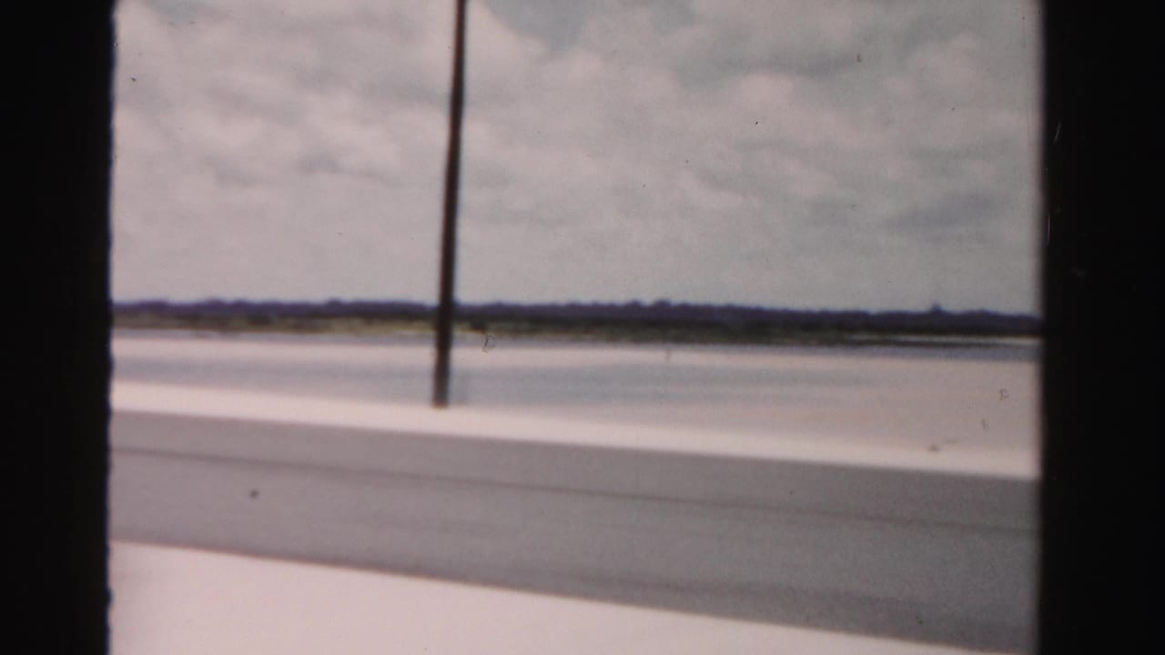1955 Dade County, Florida home-movie frame showing a bridge-side view over pale sand and water from a moving vehicle.