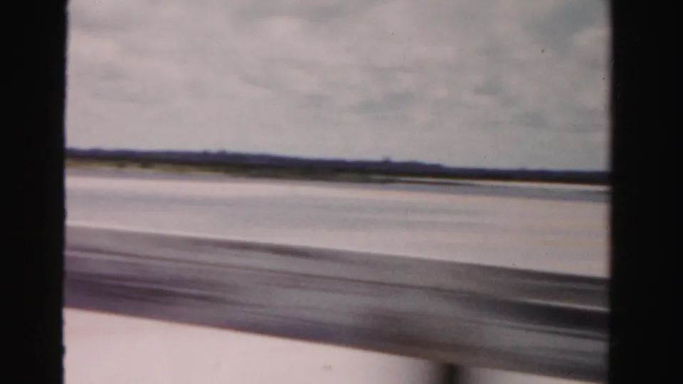 Passenger-shot 1955 Florida coastal crossing with broad sand flats, water, and a low horizon blurred by vehicle motion.