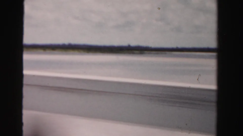 1955 Dade County home-movie frame from a moving car, looking past the window toward water and a distant low shoreline.
