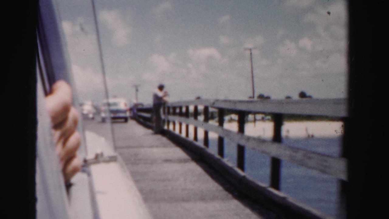 Hand on a car window as the vehicle passes a wooden railing by water.
