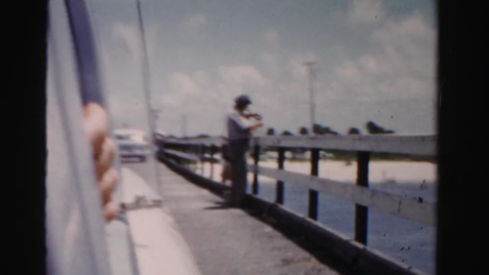 From a moving car, the frame shows a hand at the window, a fisherman near a wooden coastal railing, cars, water, and open sky.