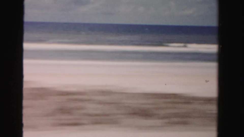 Motion-blurred 1955 Dade County coastal view of sandy beach flats, ocean water, horizon, and cloudy sky from a moving vehicle.