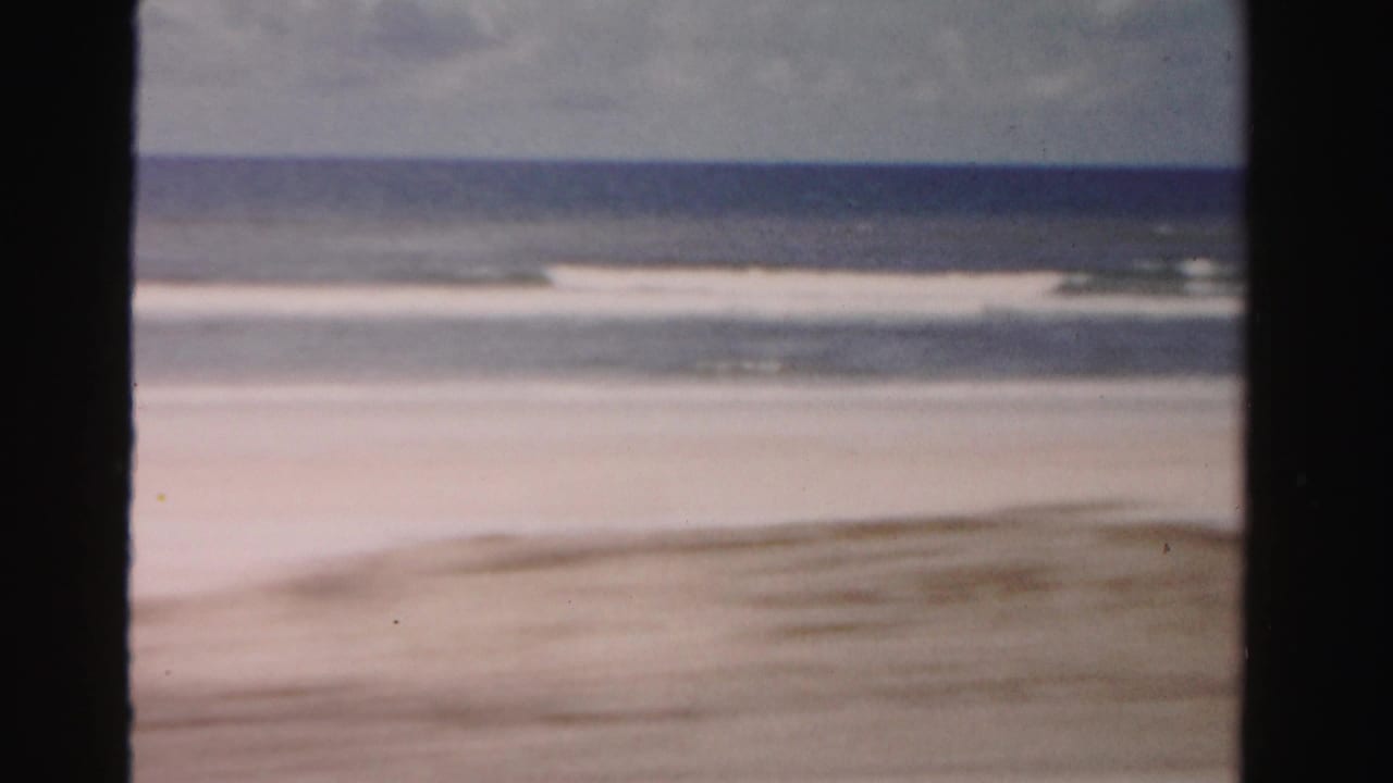 Soft surf reaches a wide sandy beach under a cloudy sky, framed by dark film borders.