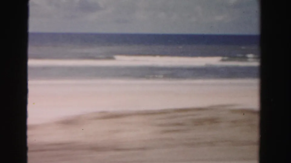 1955 Dade County 8mm frame showing pale sand, low ocean waves, overcast sky, and dark film-gate edges.