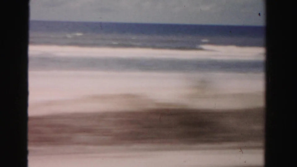 1955 Dade County 8mm frame with motion-blurred beach foreground, ocean surf, cloudy sky, and film border.
