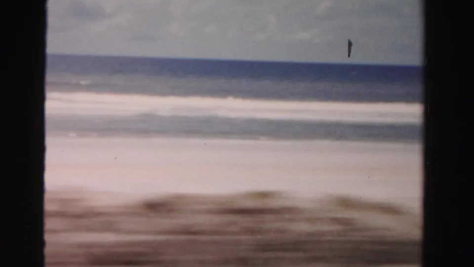 1955 Dade County shoreline frame showing pale beach, low surf, cloudy horizon, and visible film-gate edges.