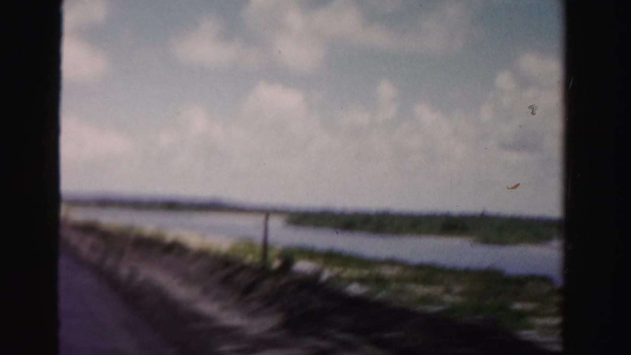 1955 Dade County 8mm frame looking from a moving vehicle past a dark window edge toward water, sand, vegetation, and sky.