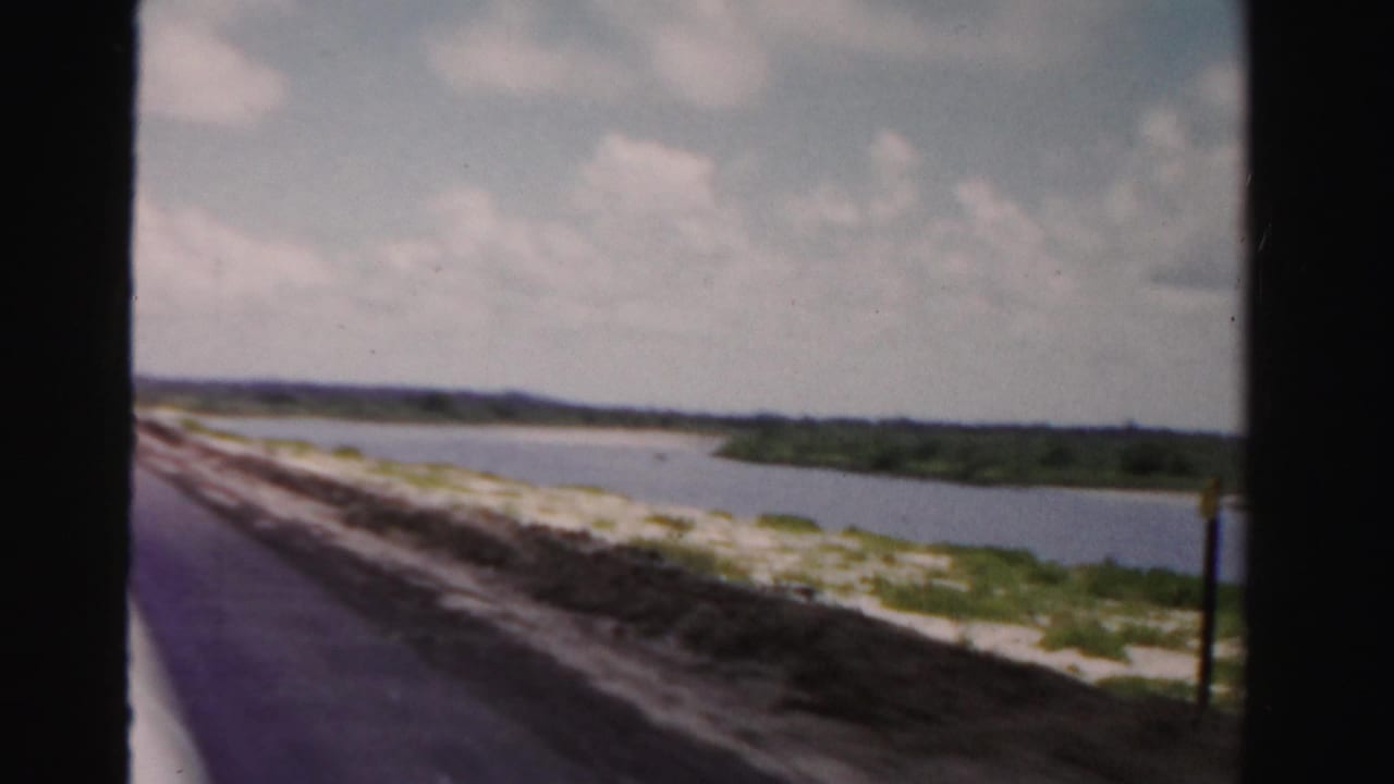 Roadside view along water with a dark window divider, sandy shoulder, low vegetation, and clouds.