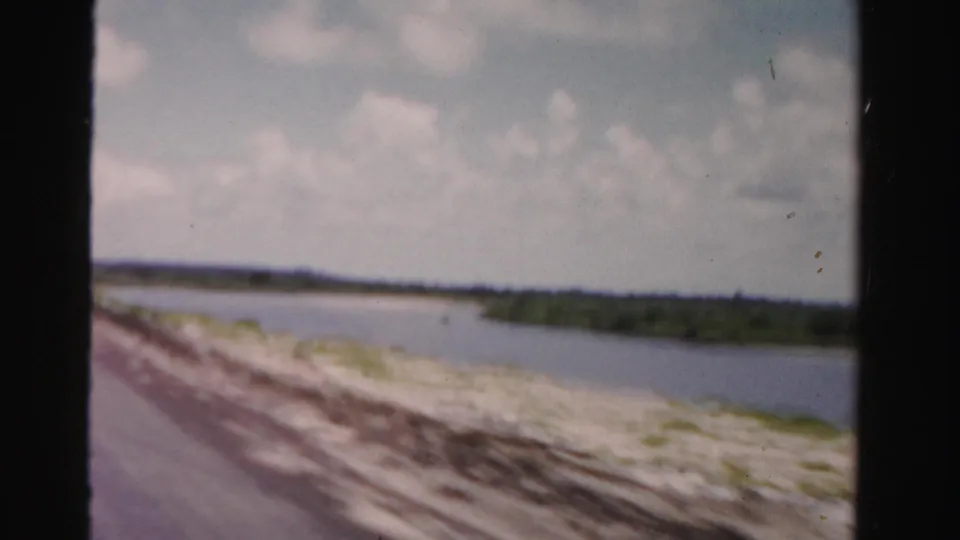The 1955 home-movie frame shows a blurred roadside sweep past sand and water with low green land in the distance.