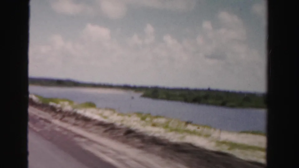 Dade County 8mm footage frames a waterway and sandy bank through a vehicle window, with low vegetation and clouds beyond.