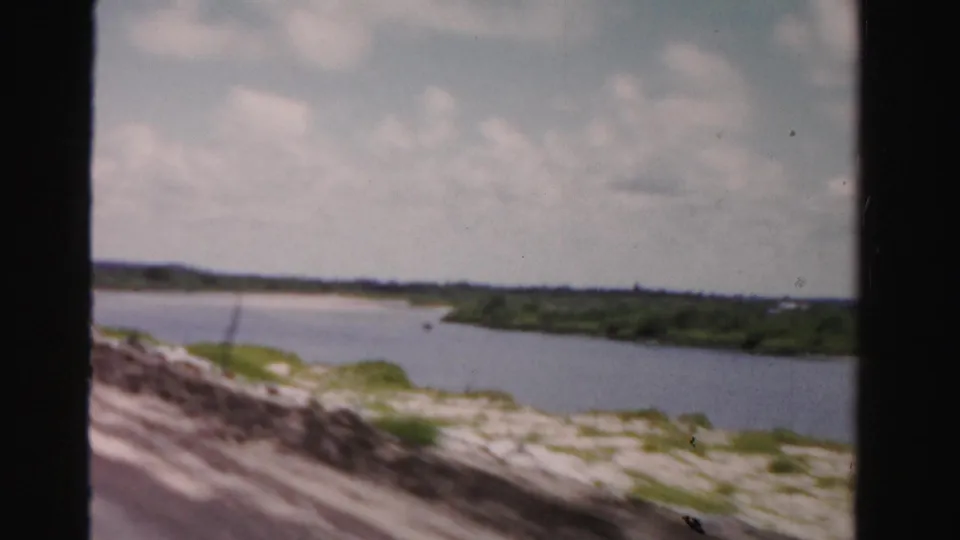 1955 Dade County, Florida 8mm frame showing a waterway, low green shoreline, blurred roadside foreground, and partly cloudy sky.