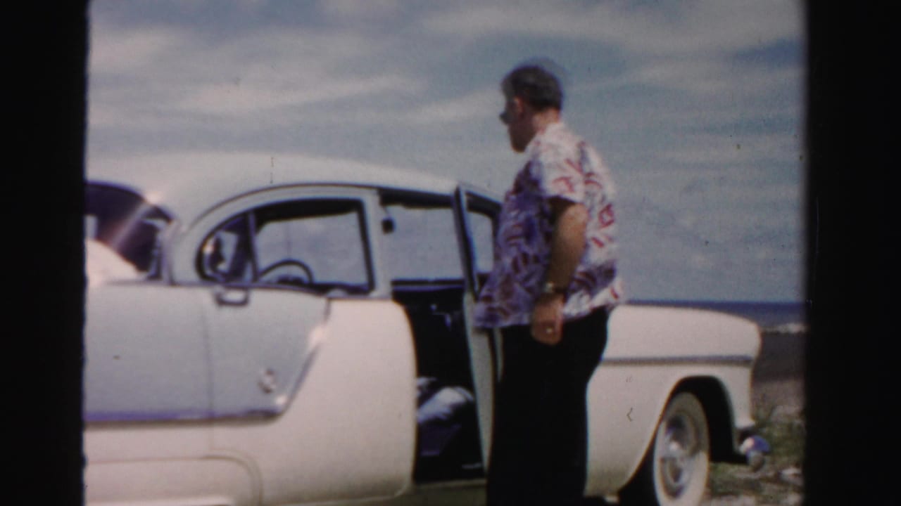 Adult man in sunglasses stands by a white and blue 1950s car near water in Dade County, Florida, with the driver's door open.