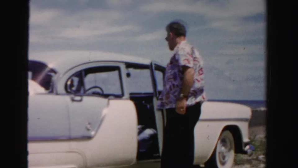 Adult man in sunglasses stands by a white and blue 1950s car near water in Dade County, Florida, with the driver's door open.
