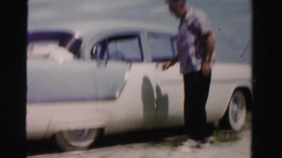 Motion-blurred 8mm frame of the man beside the white and blue car as he moves near the driver's side in 1955 Dade County footage.