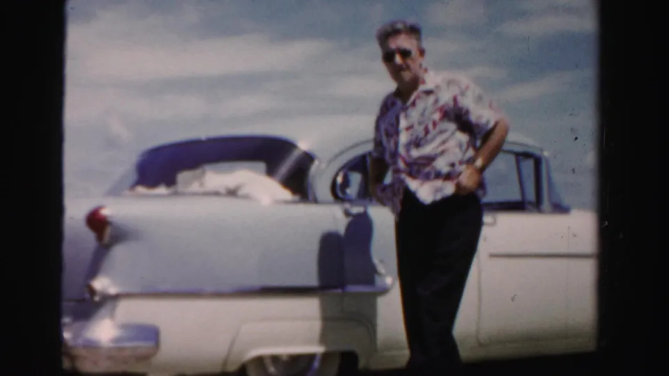 Adult man in a patterned shirt and sunglasses stands beside a white and blue 1950s automobile near open sky and water in Dade County, Florida.