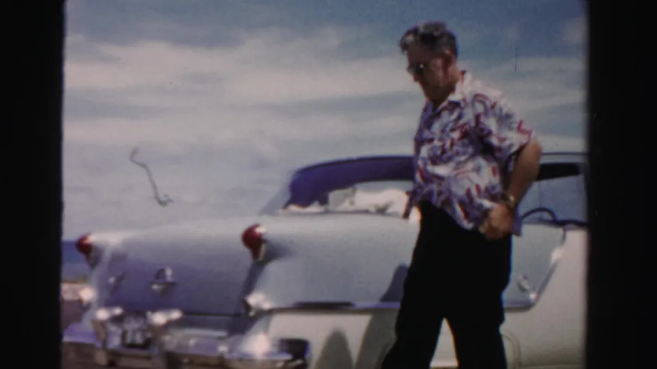 In 1955 Dade County, Florida footage, a man in sunglasses and patterned shirt stands by a light-colored car on a coastal roadside.