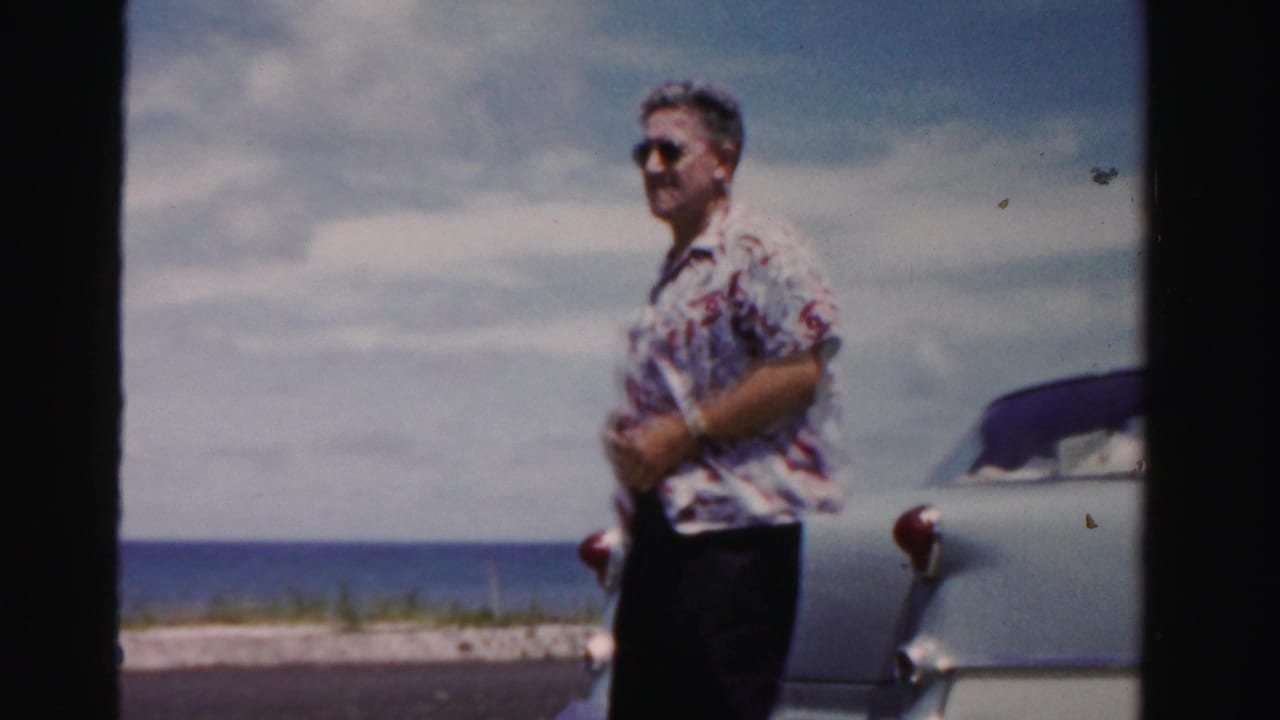 Man in sunglasses stands near the rear of a light-colored car by the coast.