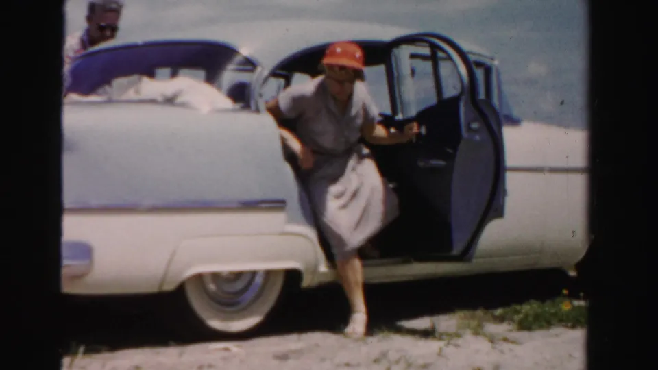 In Dade County, Florida, a woman in a red hat and light dress exits a blue and white 1950s sedan on sandy ground.