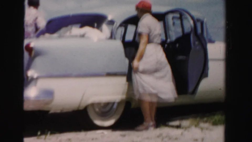 A woman in a red hat and light dress stands near the open door of a blue and white sedan in 1955 Dade County footage.