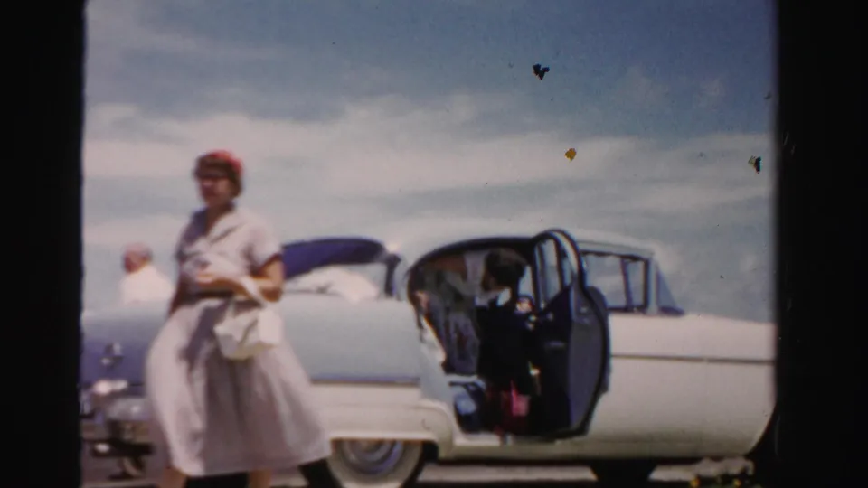 The woman stands near the front of the two-tone sedan as an open rear door reveals a seated figure inside.