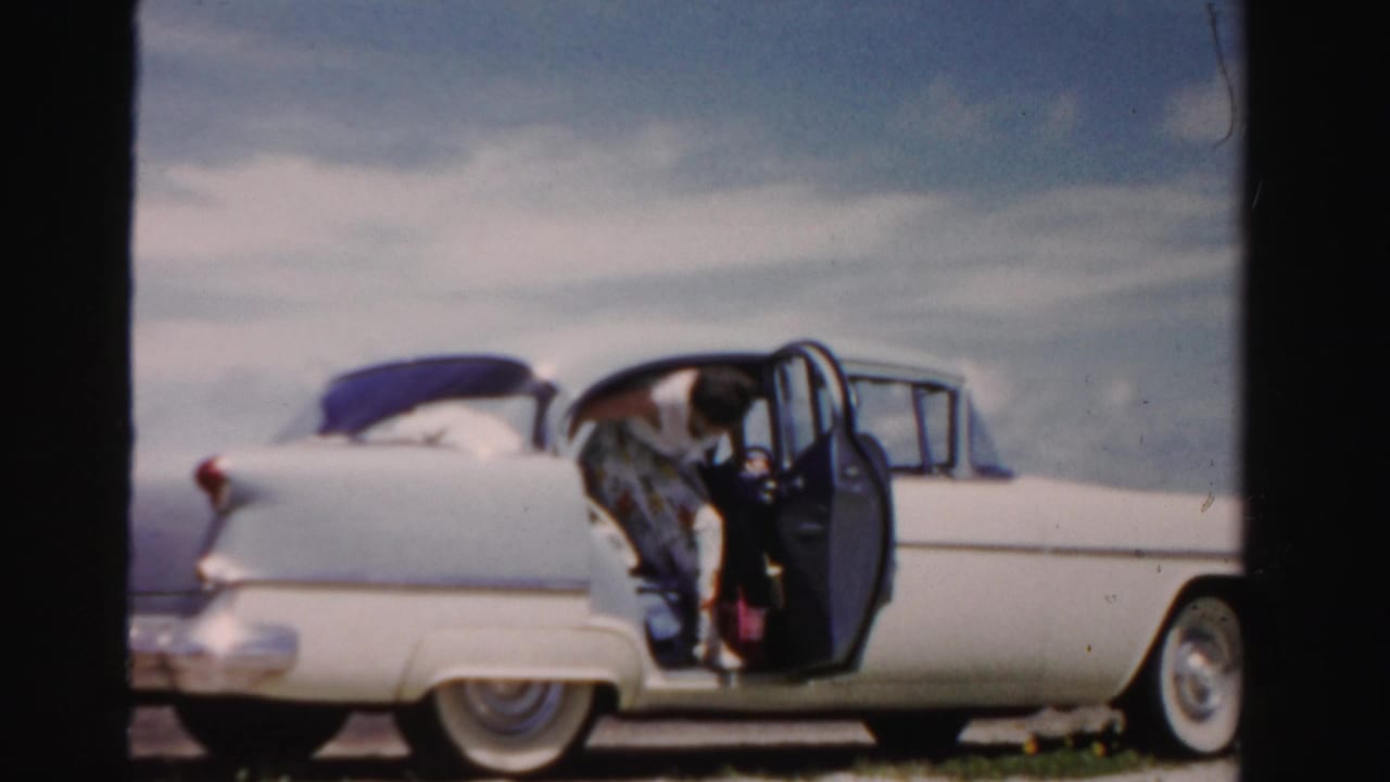 Woman partly inside the open rear door of a white vintage sedan under a bright sky.