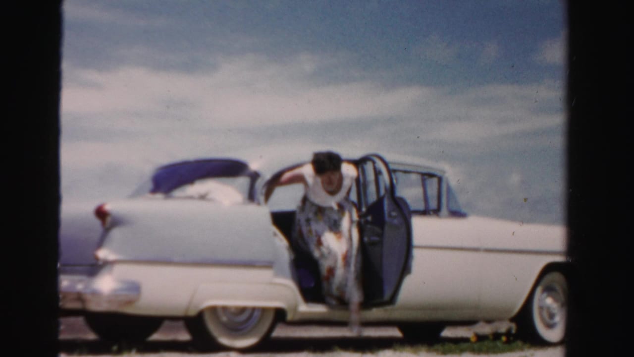 The woman leans forward from the open rear door of the white sedan, with the car's tail fin and rounded rear body visible in the frame.