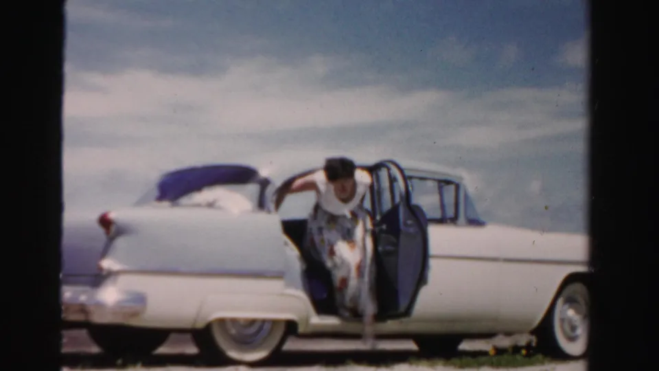 The woman leans forward from the open rear door of the white sedan, with the car's tail fin and rounded rear body visible in the frame.