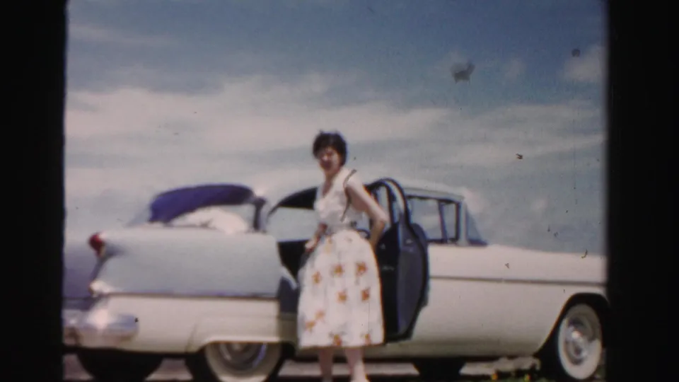 The woman stands beside the open rear door in a light floral dress, with the white 1950s sedan and bright outdoor setting behind her.