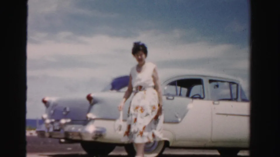 The woman walks in front of the white sedan carrying a purse, with the rear fender, chrome details, and open-air roadside setting visible.