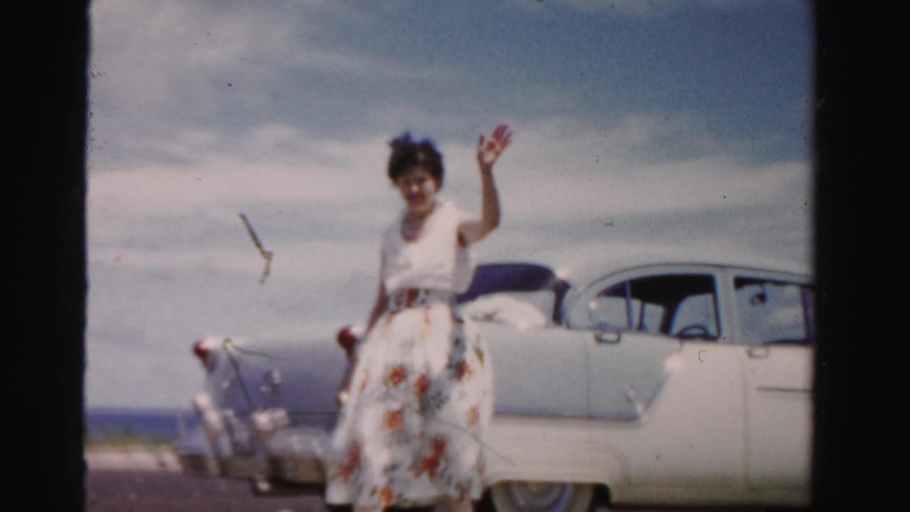 Woman waves beside a light two-tone car near the beach.