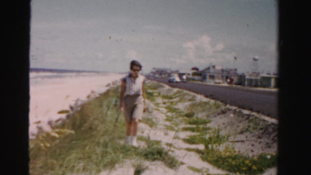 Woman walks along a sandy path beside a beach road.