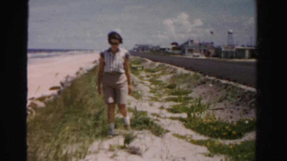 Adult woman in shorts and sunglasses walks toward the camera along a beach dune path with ocean and roadside buildings visible.