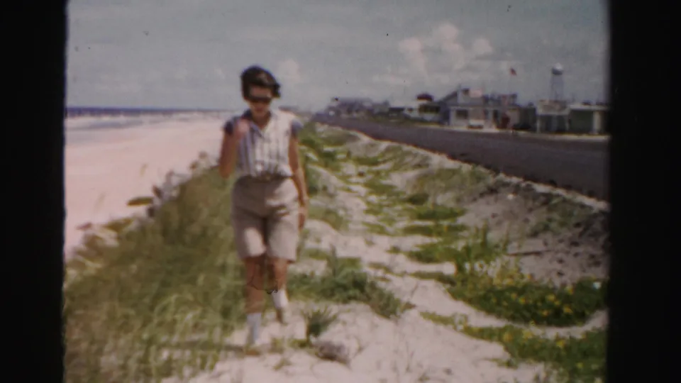 In the final frame, the woman walks closer along the dune path between the beach and paved road in 1950s Dade County footage.