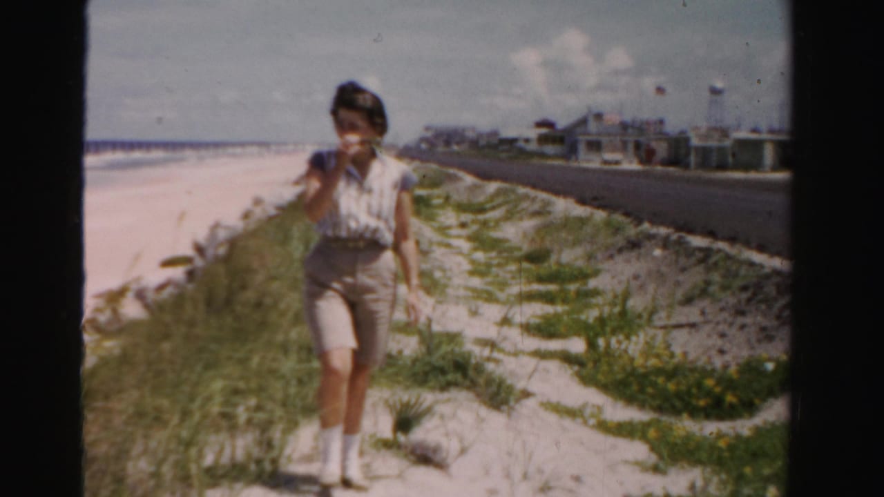 Woman walking on a sandy beachside path beside dunes and a paved road.