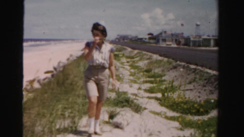In 1955 Dade County, Florida, a woman walks toward the camera along a sandy path between beach grass and an oceanfront road.