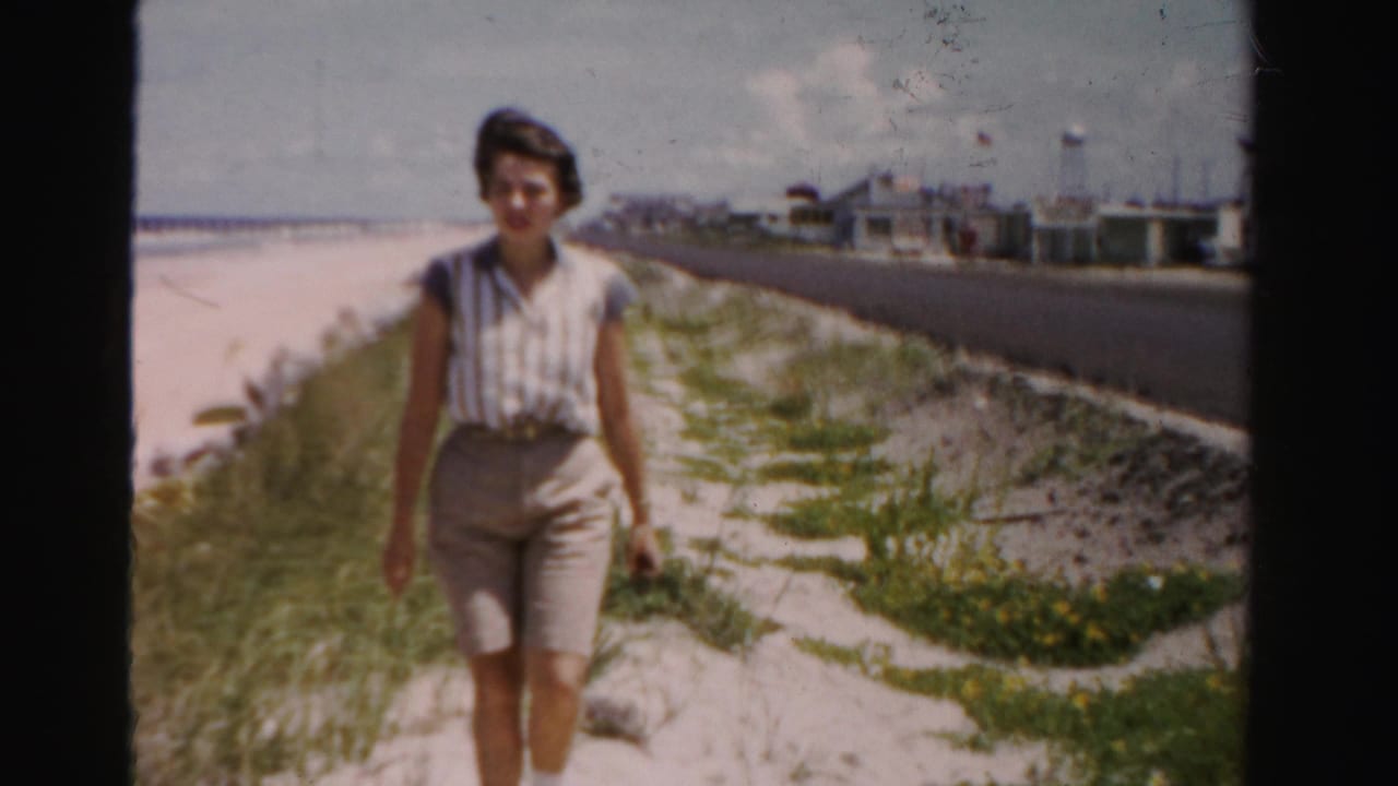 A woman in a sleeveless striped blouse and light shorts walks along a Dade County beachside path with dunes, road, and distant structures behind her.