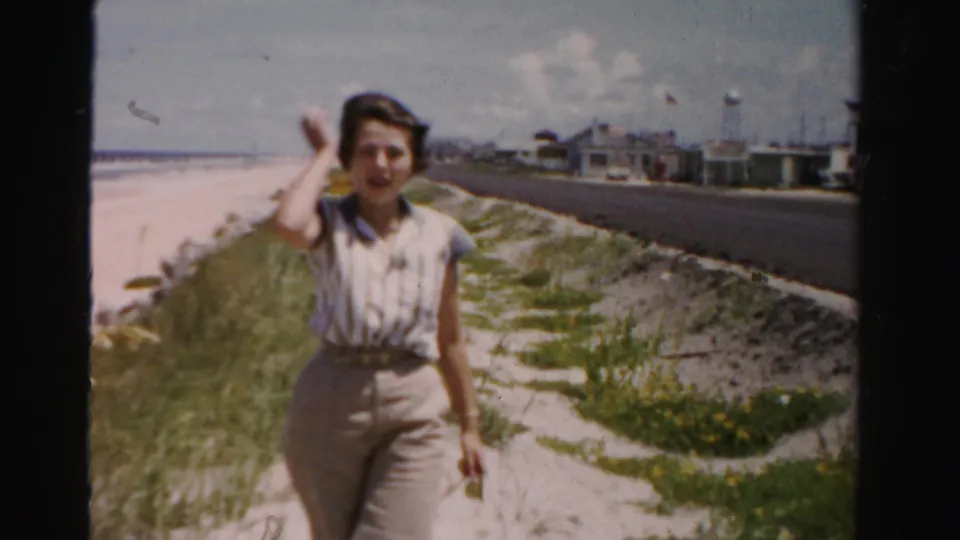 The woman raises a hand toward the camera while walking on the sandy shoreline path in 1955 Dade County, with road and beach plants visible.