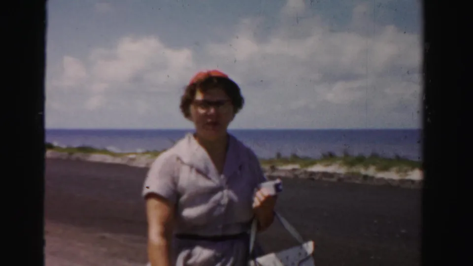 A second woman wearing a red hat and glasses stands near the beachside road, holding a small object with the ocean and clouds in the background.