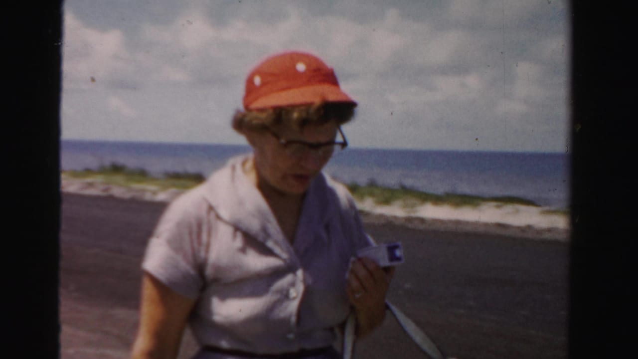 Woman in a red sun hat walks beside a coastal road with ocean behind her.