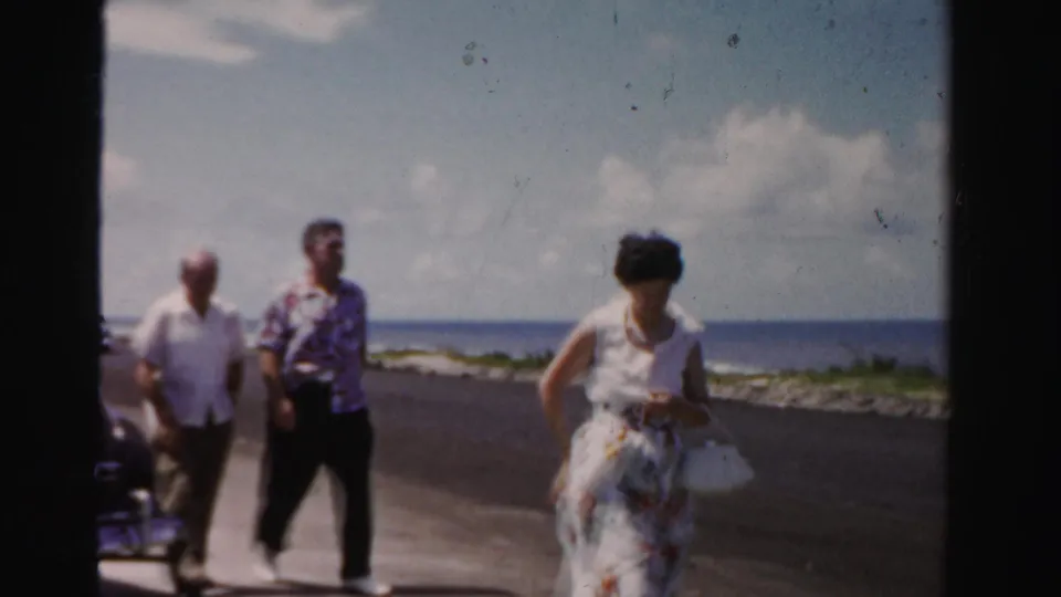 Three adults walk along a Dade County beachside road, including a woman in a floral dress and a man in a patterned shirt.