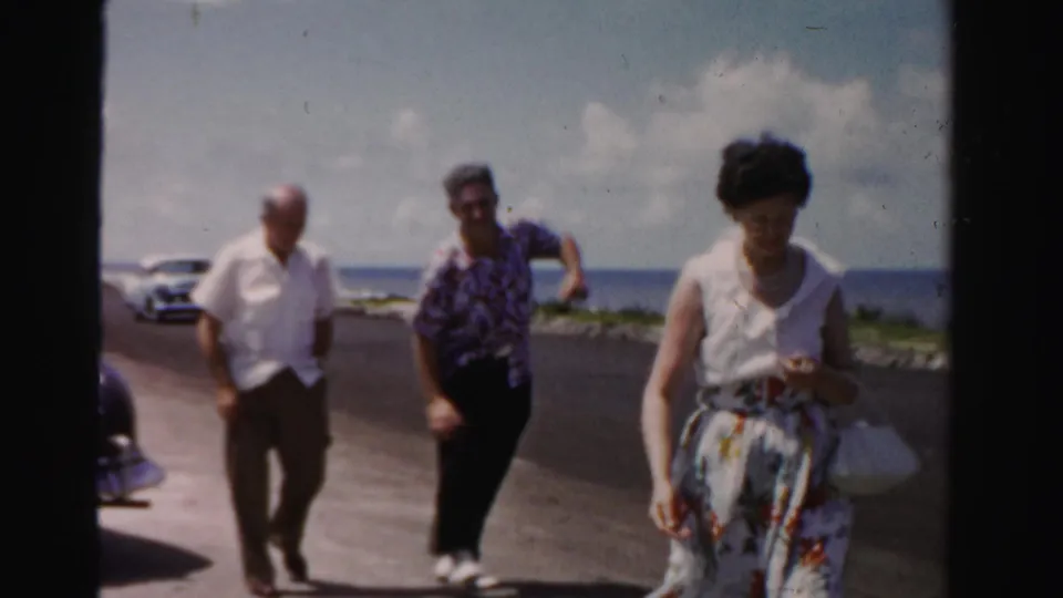 Adults pass a parked car on a Florida coastal road, with a woman in a floral skirt closest to the camera and ocean beyond.